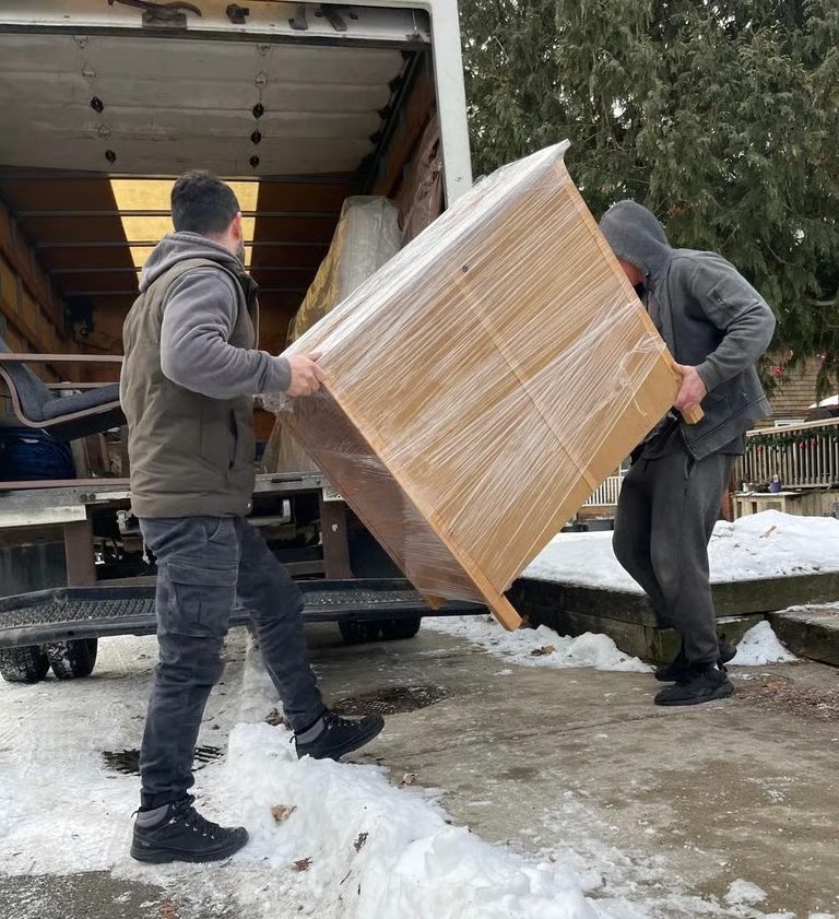 Two movers loading a shrink-wrapped dresser into a truck on a snowy Calgary driveway